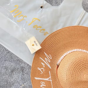 May include: A clear tote bag with gold lettering that reads "Mrs. Tocco" is next to a straw sun hat with the same text in white. A small cream-colored pouch with gold hardware is also present.
