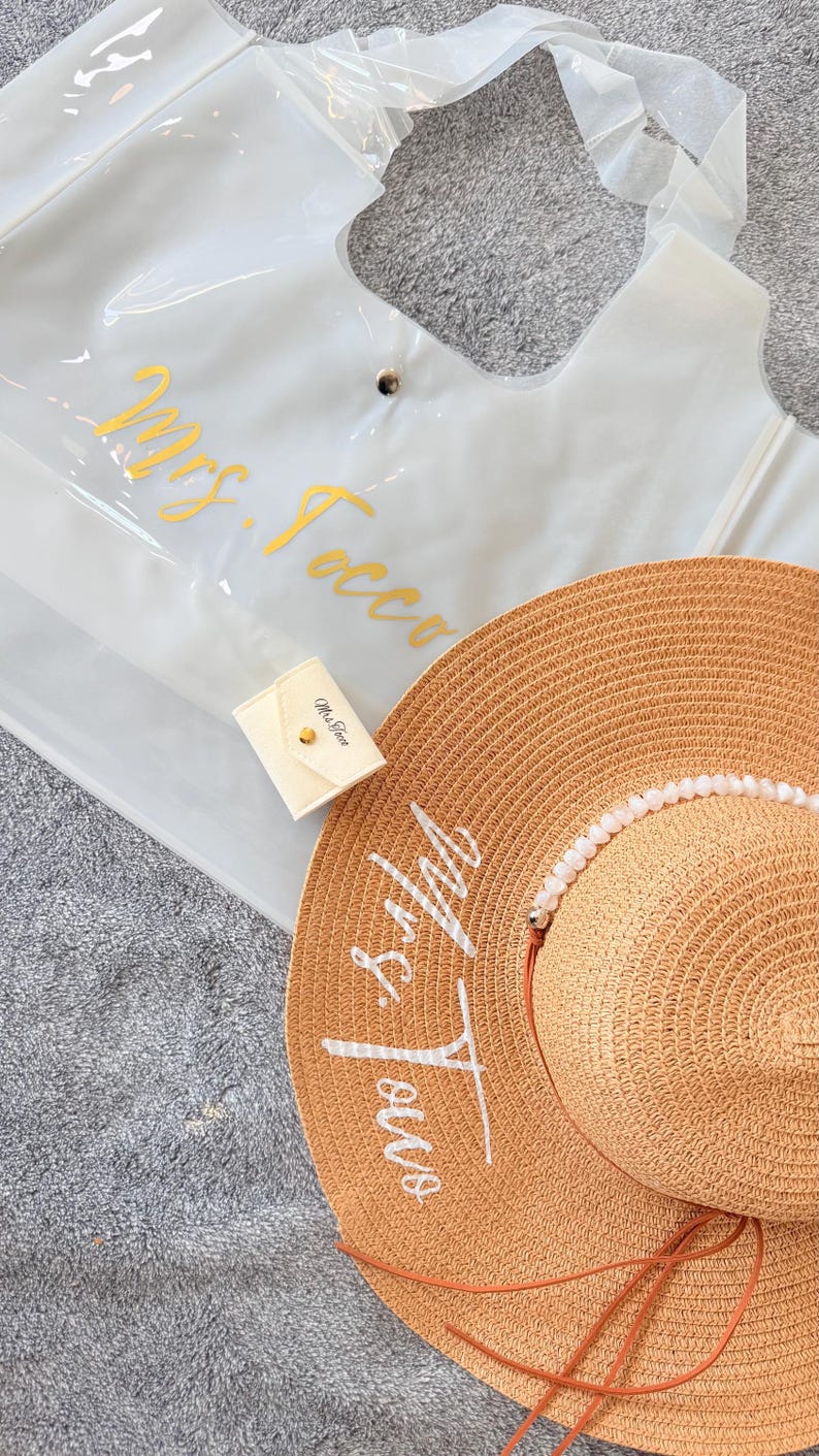 May include: A clear tote bag with gold lettering that reads "Mrs. Tocco" is next to a straw sun hat with the same text in white. A small cream-colored pouch with gold hardware is also present.