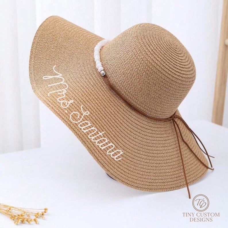 May include: A woven straw sun hat with a wide brim and a rounded crown. The hat features the embroidered text "Mrs Santana" in white script. A decorative band with white beads and a brown tie detail the hat.