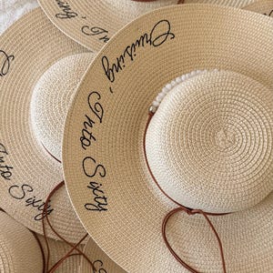 May include: A collection of beige straw sun hats with wide brims. Each hat features a smaller, rounded crown and a brown leatherette band. The phrase "Cruising Into Sixty" is embroidered in black script on the brim.