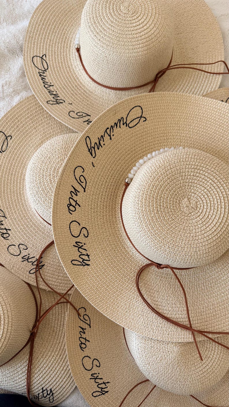 May include: A collection of beige straw sun hats with wide brims. Each hat features a smaller, rounded crown and a brown leatherette band. The phrase "Cruising Into Sixty" is embroidered in black script on the brim.