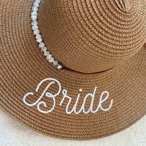 May include: A wide-brimmed, woven sun hat in a light brown hue. The hat features a white beaded band and the word "Bride" embroidered in white script across the brim. Brown ties hang from the hat.