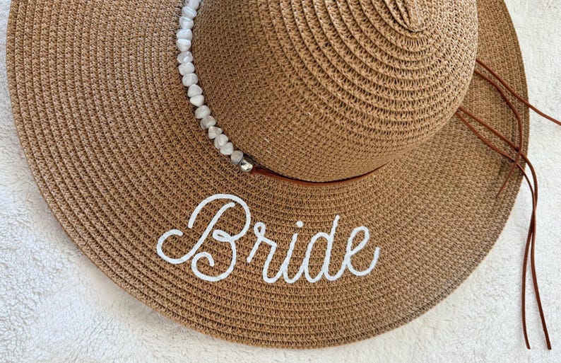May include: A wide-brimmed, woven sun hat in a light brown hue. The hat features a white beaded band and the word "Bride" embroidered in white script across the brim. Brown ties hang from the hat.