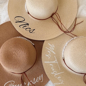May include: Three straw sun hats in varying shades of beige and brown. Each hat features a wide brim and a smaller, rounded crown. The hats are adorned with text in cursive script, and have a brown string detail. One hat says "Nics", another says "Guernsey", and the third says "Mrs. Tringley".