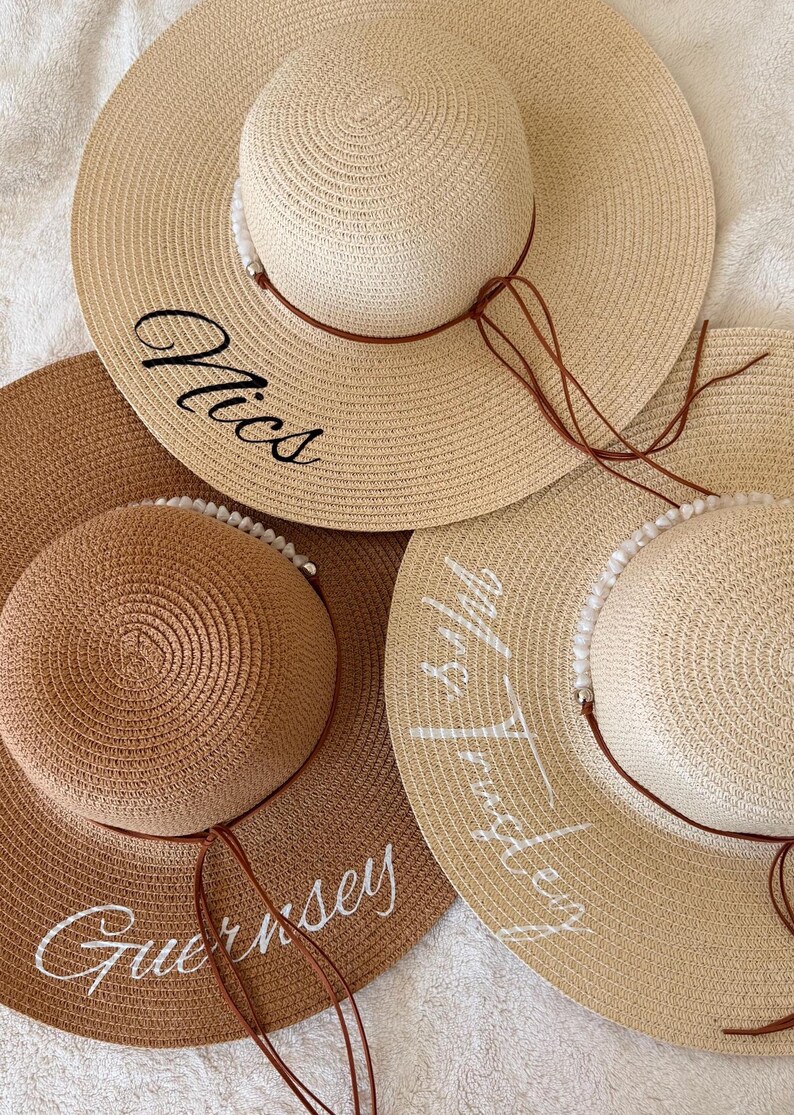 May include: Three straw sun hats in varying shades of beige and brown. Each hat features a wide brim and a smaller, rounded crown. The hats are adorned with text in cursive script, and have a brown string detail. One hat says "Nics", another says "Guernsey", and the third says "Mrs. Tringley".