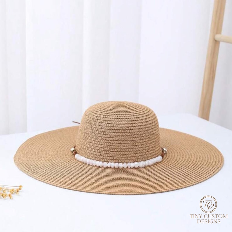 May include: A wide-brimmed, tan straw hat with a rounded crown. The hat features a decorative band of white beads around the base of the crown. The hat is designed for sun protection and is suitable for outdoor wear. The logo "TINY CUSTOM DESIGNS" is visible.