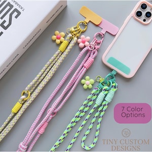 May include: A clear phone case with a pink border is attached to a lanyard with a yellow, pink, and green rope design. The lanyards have flower charms and are labeled with "7 Color Options" and "Tiny Custom Designs".
