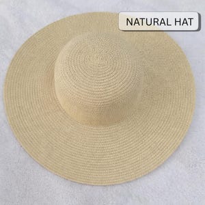 May include: A wide-brimmed, natural-colored straw hat. The hat features a rounded crown and a broad, circular brim. The text "NATURAL HAT" is displayed on a white rectangular label. The hat is made of woven straw material.