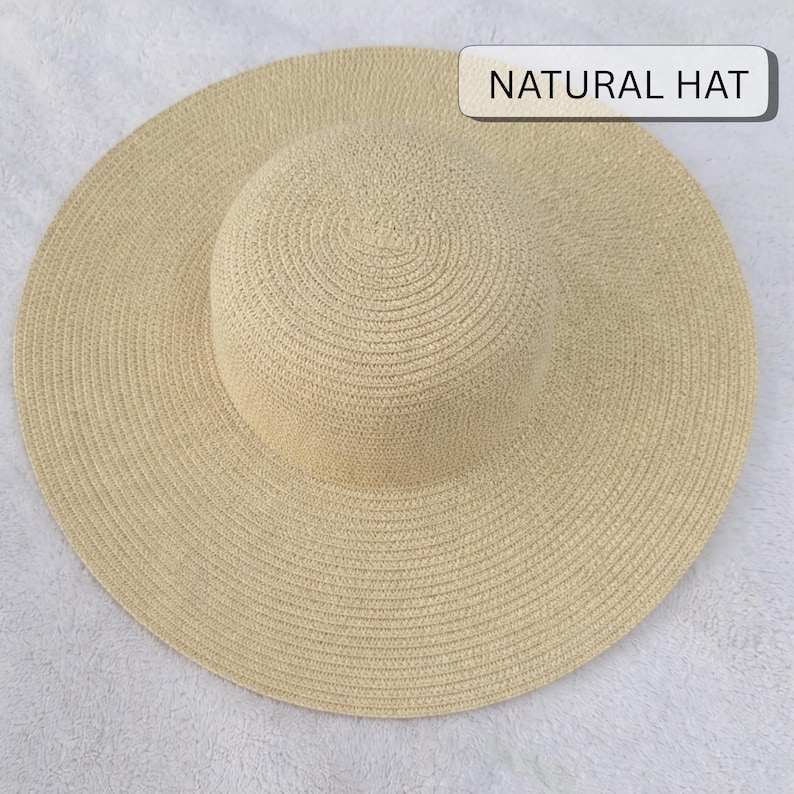 May include: A wide-brimmed, natural-colored straw hat. The hat features a rounded crown and a broad, circular brim. The text "NATURAL HAT" is displayed on a white rectangular label. The hat is made of woven straw material.
