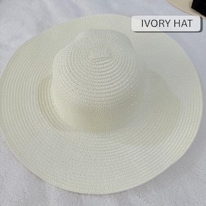 May include: An ivory-colored sun hat with a wide brim. The hat has a rounded crown and a broad, circular brim. The text "IVORY HAT" is displayed on a white rectangular label. The hat is made of woven material.