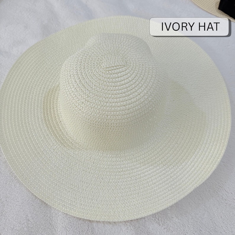 May include: An ivory-colored sun hat with a wide brim. The hat has a rounded crown and a broad, circular brim. The text "IVORY HAT" is displayed on a white rectangular label. The hat is made of woven material.