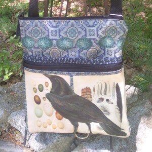 May include: A black crossbody bag with a zippered closure. The bag features a patterned fabric with blue, green, and gold accents. The front of the bag has a print of a black raven and a collection of eggs and feathers.