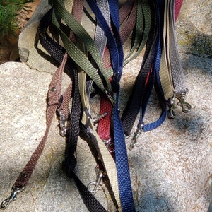 May include: A collection of 12 colorful woven straps with metal clasps. The straps are arranged in a pile on a gray rock surface.