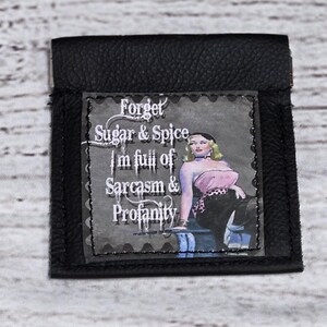 May include: Black leather wallet with a fabric patch that says "Forget Sugar & Spice I'm full of Sarcasm & Profanity". The patch features a vintage style illustration of a woman in a pink and black outfit.