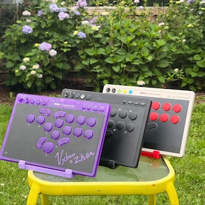 Könnte beinhalten: Drei rechteckige Arcade-Controller in einer Reihe. Der vordere Controller ist durchscheinend lila mit lila Tasten und dem Text Vxbao & Zhen. Der mittlere Controller ist schwarz mit schwarzen Tasten. Der hintere Controller ist beige mit roten Tasten.