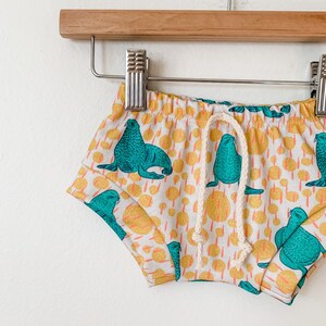 May include: White baby shorts with a drawstring waist and a teal and yellow print of sea lions.