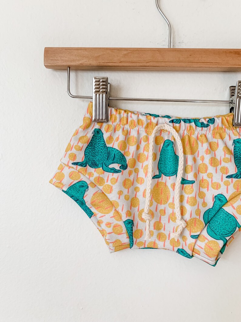 May include: White baby shorts with a drawstring waist and a teal and yellow print of sea lions.