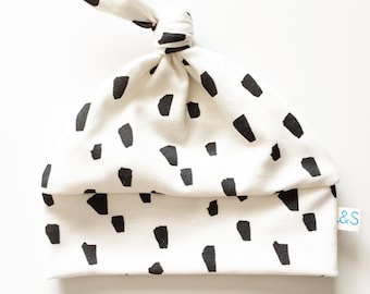 May include: White baby hat with a black and white abstract print. The hat has a knot on top and a small tag that says "&S".