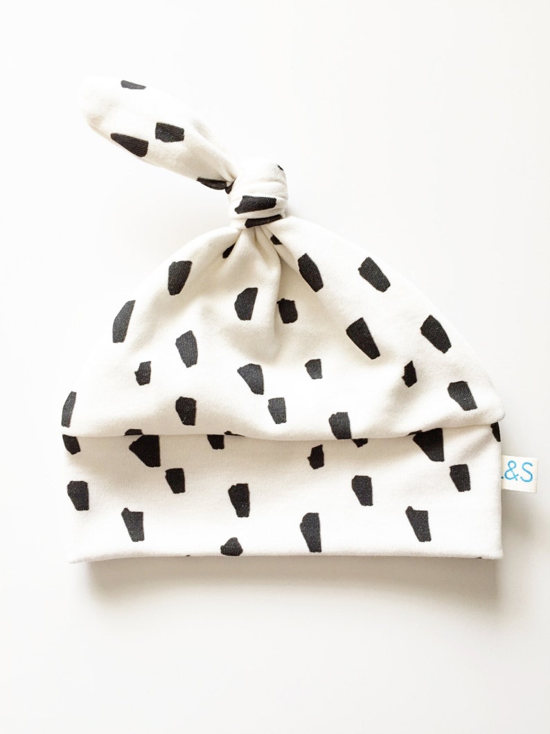 May include: White baby hat with a black and white abstract print. The hat has a knot on top and a small tag that says "&S".