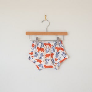 May include: White baby shorts with an orange tiger and blue leaf pattern, hanging on a wooden hanger. The shorts have a drawstring waist and are made of a soft fabric. The shorts are approximately 8 inches wide.