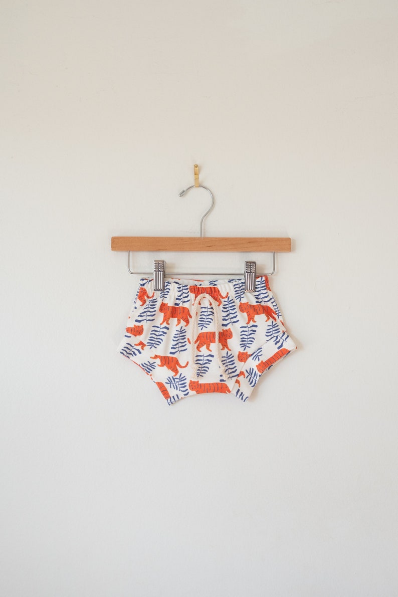 May include: White baby shorts with an orange tiger and blue leaf pattern, hanging on a wooden hanger. The shorts have a drawstring waist and are made of a soft fabric. The shorts are approximately 8 inches wide.