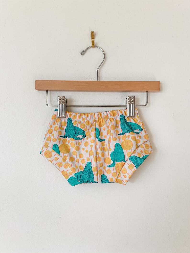 May include: White baby bloomers with a colorful print of teal sea lions on a yellow background.
