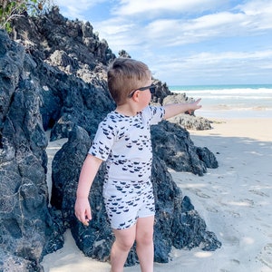 Pu&ograve; includere: Un bambino piccolo con un costume da bagno bianco e nero con un motivo a pesce si trova su una spiaggia sabbiosa, indicando l'oceano. Indossa occhiali da sole e ha i capelli corti e biondi.