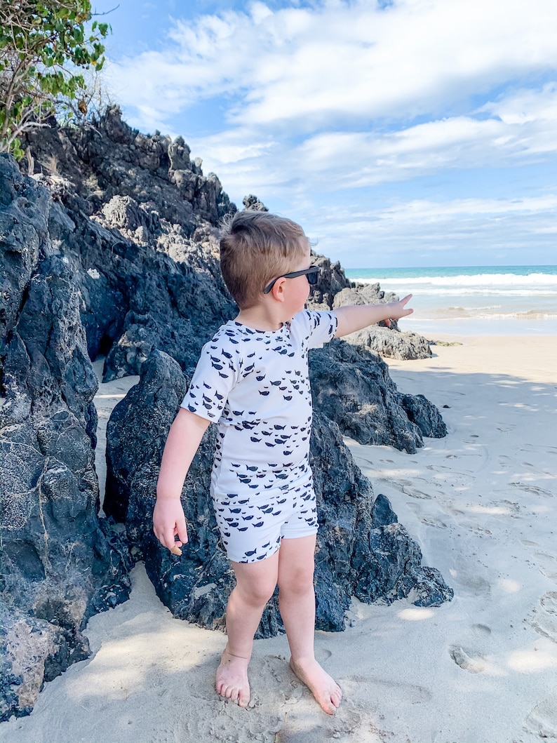 Pu&ograve; includere: Un bambino piccolo con un costume da bagno bianco e nero con un motivo a pesce si trova su una spiaggia sabbiosa, indicando l'oceano. Indossa occhiali da sole e ha i capelli corti e biondi.