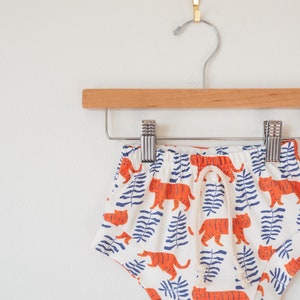 May include: White baby shorts with an orange tiger and blue leaf pattern, hanging on a wooden hanger. The shorts have a drawstring and are made of a soft fabric.