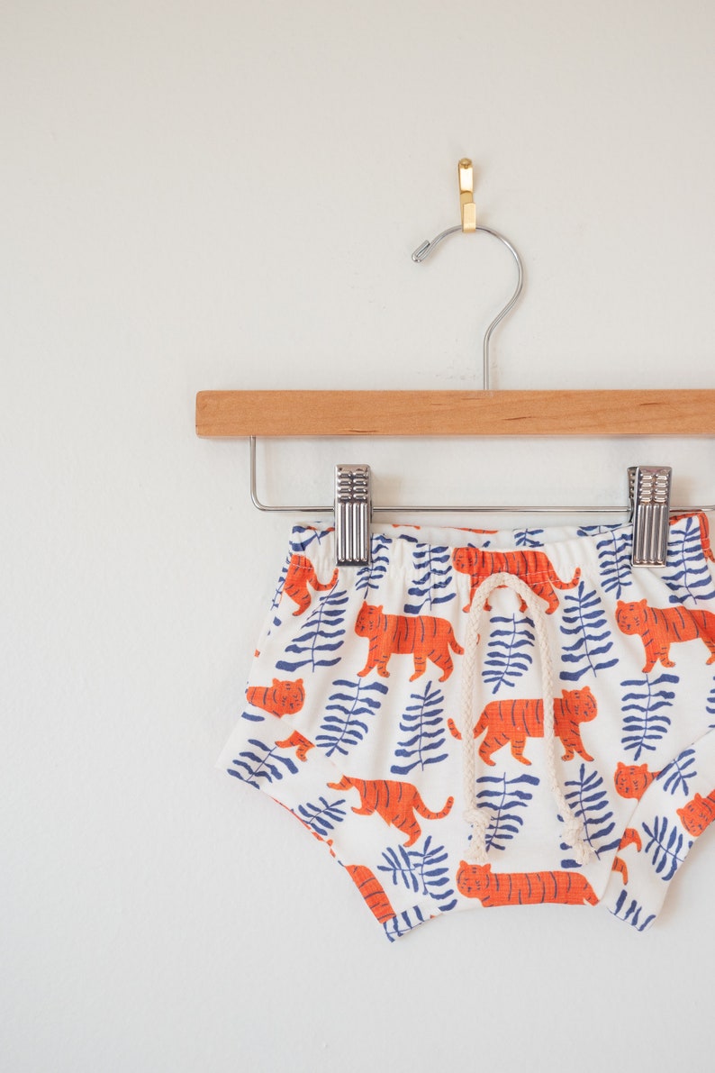 May include: White baby shorts with an orange tiger and blue leaf pattern, hanging on a wooden hanger. The shorts have a drawstring and are made of a soft fabric.