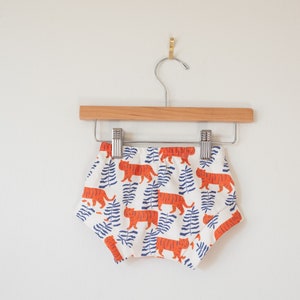 May include: White baby shorts with a repeating pattern of orange tigers and blue fern-like leaves. The shorts are hanging on a wooden hanger with a metal clip, against a white wall.