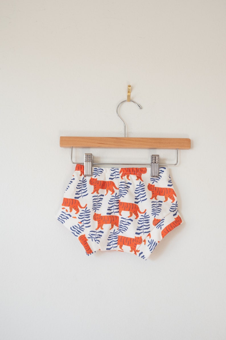 May include: White baby shorts with a repeating pattern of orange tigers and blue fern-like leaves. The shorts are hanging on a wooden hanger with a metal clip, against a white wall.