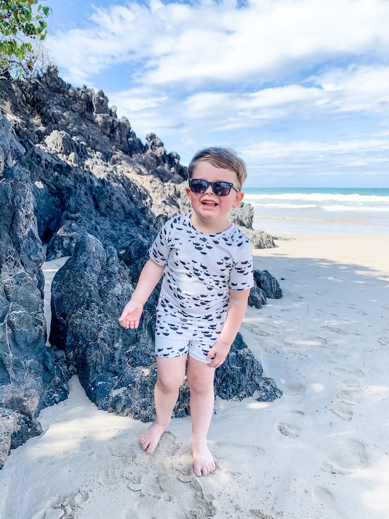 Pu&ograve; includere: Un bambino piccolo con occhiali da sole e una camicia e pantaloncini a fantasia bianca e nera sta in piedi su una spiaggia sabbiosa con rocce sullo sfondo.