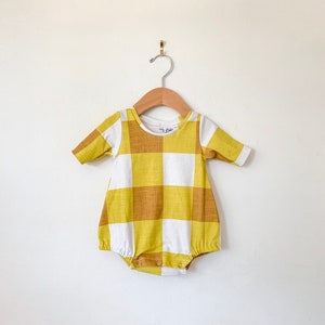 Może przedstawiać: Żółty i biały body w kratkę gingham z krótkimi rękawami. Body ma okrągły dekolt i zatrzaski na dole. Body wisi na drewnianym wieszaku.