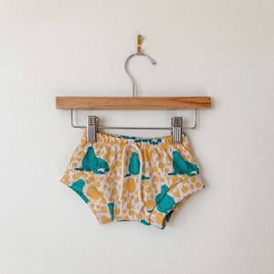 May include: White baby shorts with a yellow and teal print of sea lions. The shorts have a drawstring waist and are hanging on a wooden hanger.