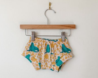 May include: White baby shorts with a yellow and teal print of sea lions. The shorts have a drawstring waist and are hanging on a wooden hanger.