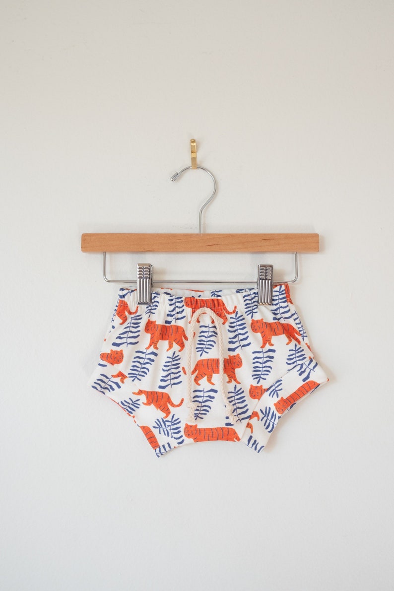 May include: White baby shorts with an orange tiger and blue leaf pattern. The shorts have a drawstring waist and are hanging on a wooden hanger. The shorts are made of a soft, lightweight fabric.
