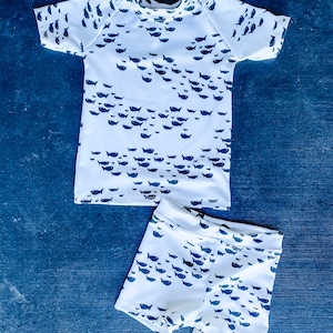 Pu&ograve; includere: Costume da bagno bianco a due pezzi per bambini con stampa di pesci blu. La parte superiore ha maniche corte e collo rotondo. I pantaloncini hanno una cintura elastica.