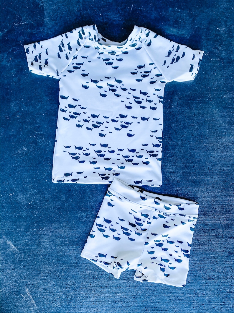 Pu&ograve; includere: Costume da bagno bianco a due pezzi per bambini con stampa di pesci blu. La parte superiore ha maniche corte e collo rotondo. I pantaloncini hanno una cintura elastica.