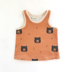 Puede incluir: Un tanque marrón sin mangas con un estampado de cara de oso negro y flechas. El tanque tiene una etiqueta blanca con el texto "Lola&Stella".