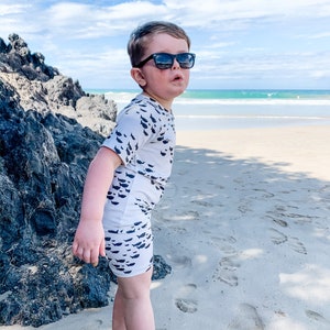 Pu&ograve; includere: Un bambino piccolo che indossa un costume da bagno bianco e nero con un motivo a pesce sta in piedi su una spiaggia sabbiosa. Il bambino indossa occhiali da sole e guarda a destra. La spiaggia &egrave; sullo sfondo con acqua blu e una costa rocciosa.