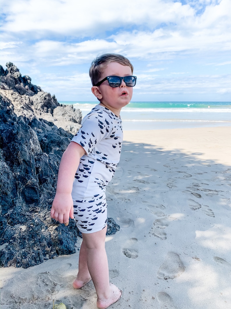 Pu&ograve; includere: Un bambino piccolo che indossa un costume da bagno bianco e nero con un motivo a pesce sta in piedi su una spiaggia sabbiosa. Il bambino indossa occhiali da sole e guarda a destra. La spiaggia &egrave; sullo sfondo con acqua blu e una costa rocciosa.
