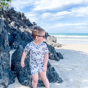 Pu&ograve; includere: Un bambino piccolo con una camicia a maniche corte bianca e nera e pantaloncini abbinati con una stampa di pesce si trova su una spiaggia sabbiosa con rocce sullo sfondo. Indossa occhiali da sole e guarda a destra.