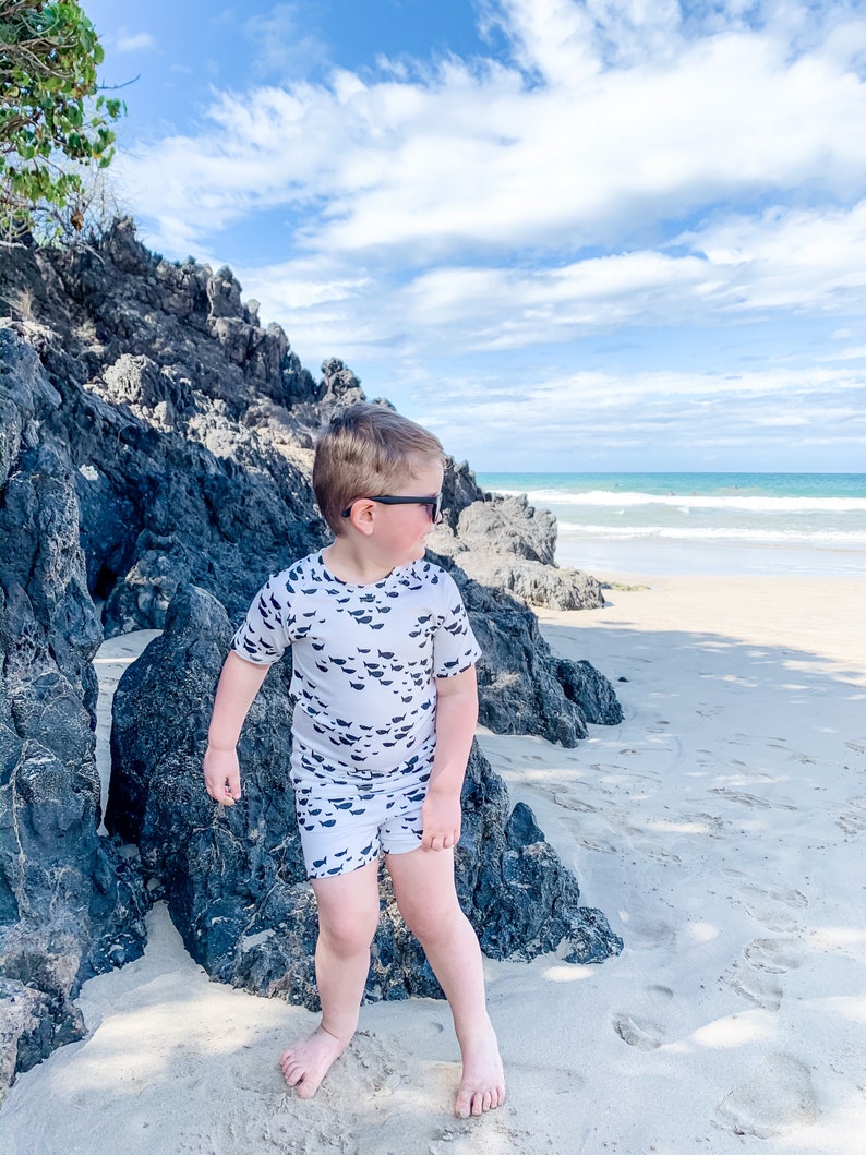 Pu&ograve; includere: Un bambino piccolo con una camicia a maniche corte bianca e nera e pantaloncini abbinati con una stampa di pesce si trova su una spiaggia sabbiosa con rocce sullo sfondo. Indossa occhiali da sole e guarda a destra.