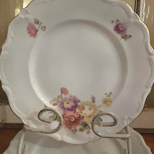 May include: White porcelain plate with a scalloped edge and gold trim. The plate features a floral design in shades of pink, purple, and yellow. The plate is displayed on a decorative metal stand.