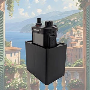 May include: A black two-way radio with the text "RORADIO" on the front, resting in a black charging cradle. The radio has an antenna and several buttons. The background is a scenic painting of a coastal town with a blue sky.