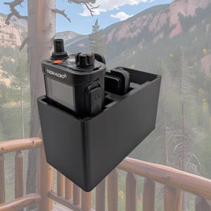 May include: Black two-way radio in a black holder. The radio has the brand name "TIDRADIO" printed on it. The holder has two slots for accessories. The background shows a mountain landscape.