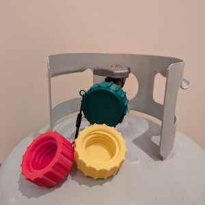May include: Three colorful propane tank caps: red, yellow, and green. The caps are made of plastic and have a ridged design. The caps are sitting on top of a propane tank.
