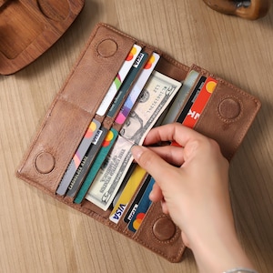 May include: A brown leather wallet with multiple card slots and a magnetic closure. The wallet is open, revealing credit cards, cash, and a hand reaching in. The wallet is on a wooden surface.