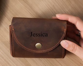 May include: A brown leather wallet with a curved flap and a brass button closure. The name "Jessica" is embossed on the front. The wallet is rectangular and appears to be handmade.